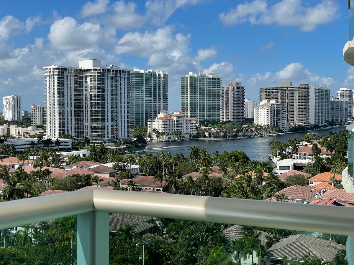 A panoramic view from the balcony showcases a vibrant skyline of tall buildings alongside a serene waterway, framed by lush greenery. The scene captures a mix of urban life and natural beauty, inviting moments of relaxation and enjoyment.