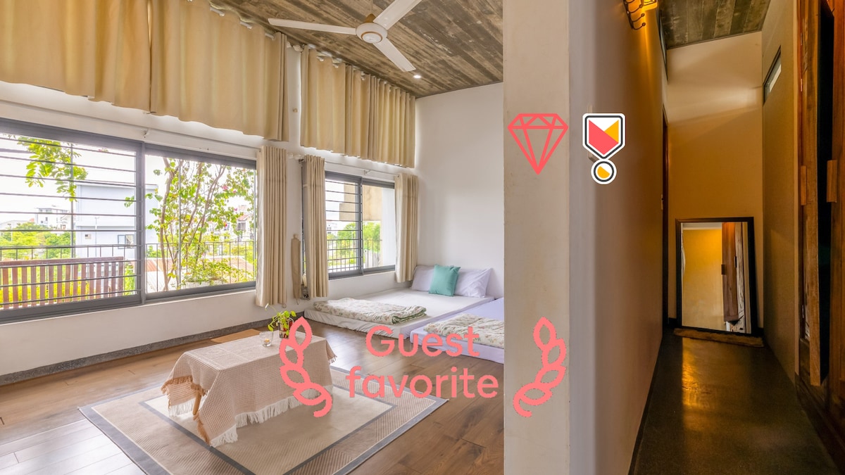 A bright and airy studio space features large windows that invite natural light. A minimalistic layout includes a cozy sleeping area with soft bedding and a calm color palette. A simple rug is placed on the floor, and a hallway leads to additional areas of the home.
