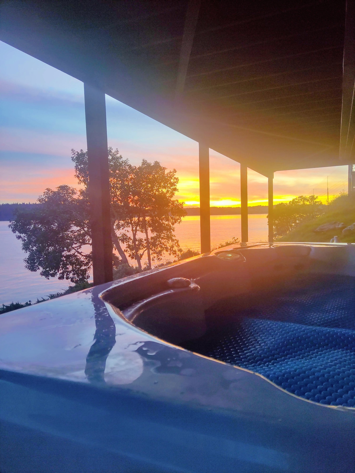 The image captures a hot tub on an outdoor deck, with tranquil waters visible nearby. A vibrant sunset illuminates the sky, casting reflections on the water's surface. Silhouetted trees frame the scene, creating a serene atmosphere for relaxation.