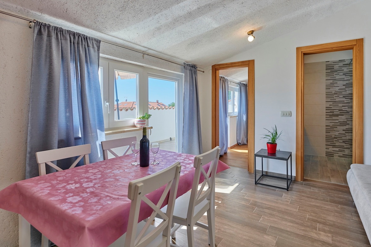 A dining area is presented with a pink tablecloth draped over a white table surrounded by four wooden chairs. Large windows allow natural light to fill the space, while light blue curtains frame the glass. An entrance to a room is visible in the background.