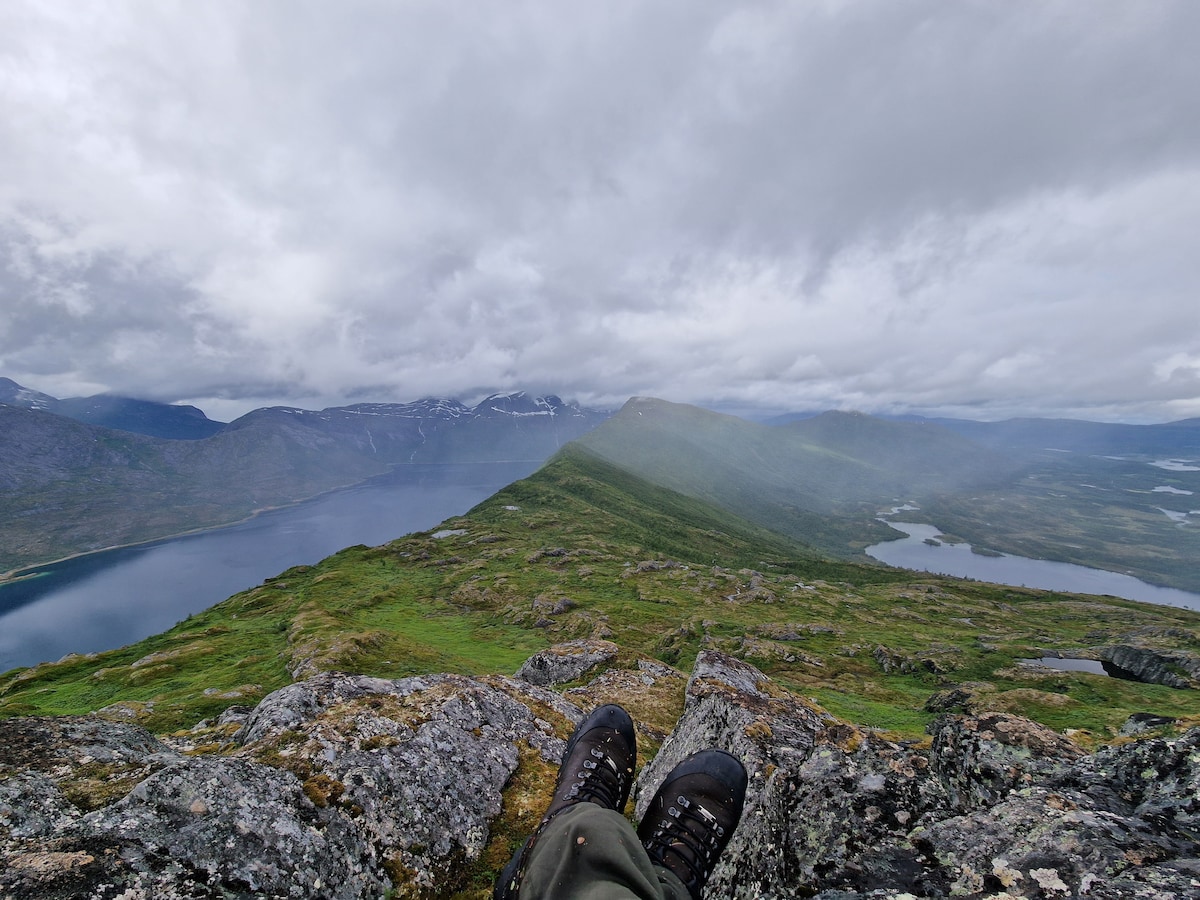 A panoramic view from a rocky vantage point is presented, showcasing lush green hills sloping down to a serene lake. Dark clouds hover above, creating a dramatic sky against the distant mountains, enhancing the natural beauty of the Norwegian landscape.