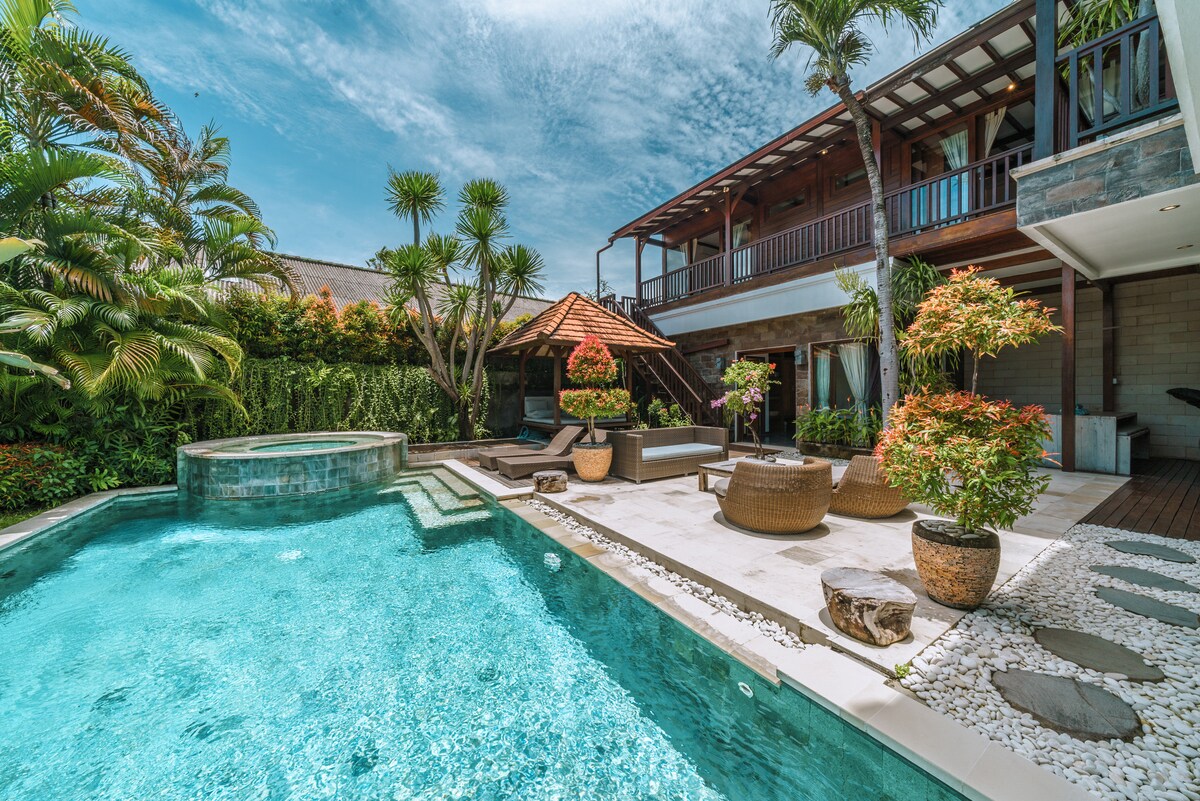 A large swimming pool is surrounded by lush tropical plants and stone pathways. Woven lounge chairs are positioned near the water, alongside potted plants that add vibrant colors. A spacious deck leads to a second-level patio with a traditional roof structure, providing shaded seating areas.