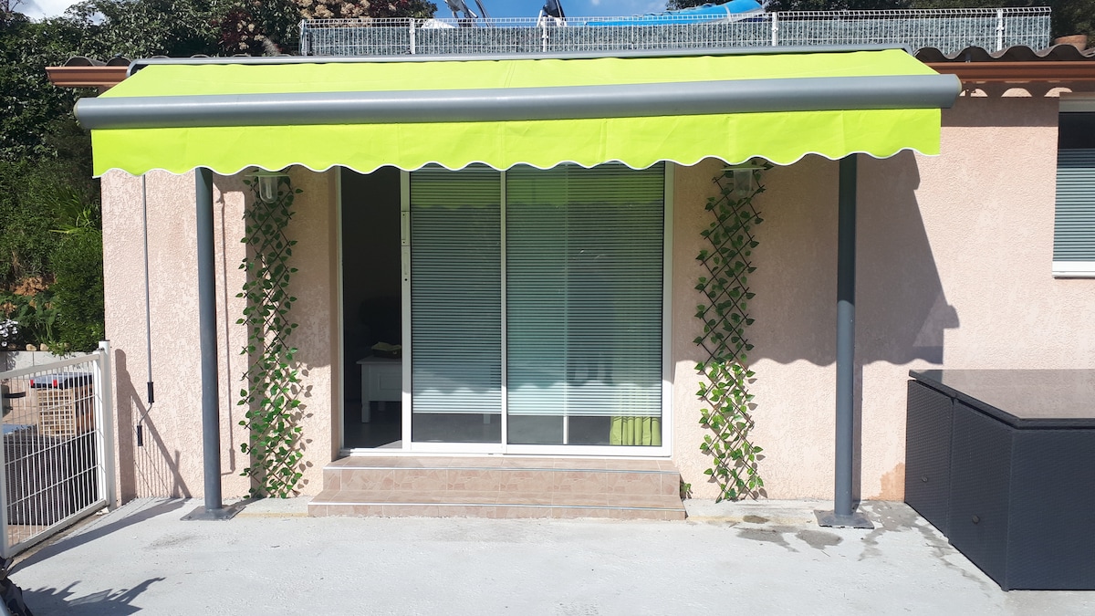 A covered entryway features a bright green awning and is flanked by decorative climbing plants. Sliding glass doors provide a view into the living area, complemented by a light-colored exterior wall. A small step leads to the door in a clean pavement area.
