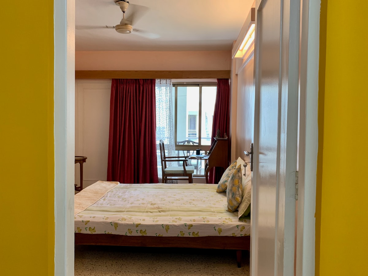 A well-lit room is viewed through an open door, featuring a spacious bed adorned with light-colored linens and decorative pillows. Nearby, a window with sheer curtains reveals a small seating area with a table and chairs, complemented by rich red curtains.