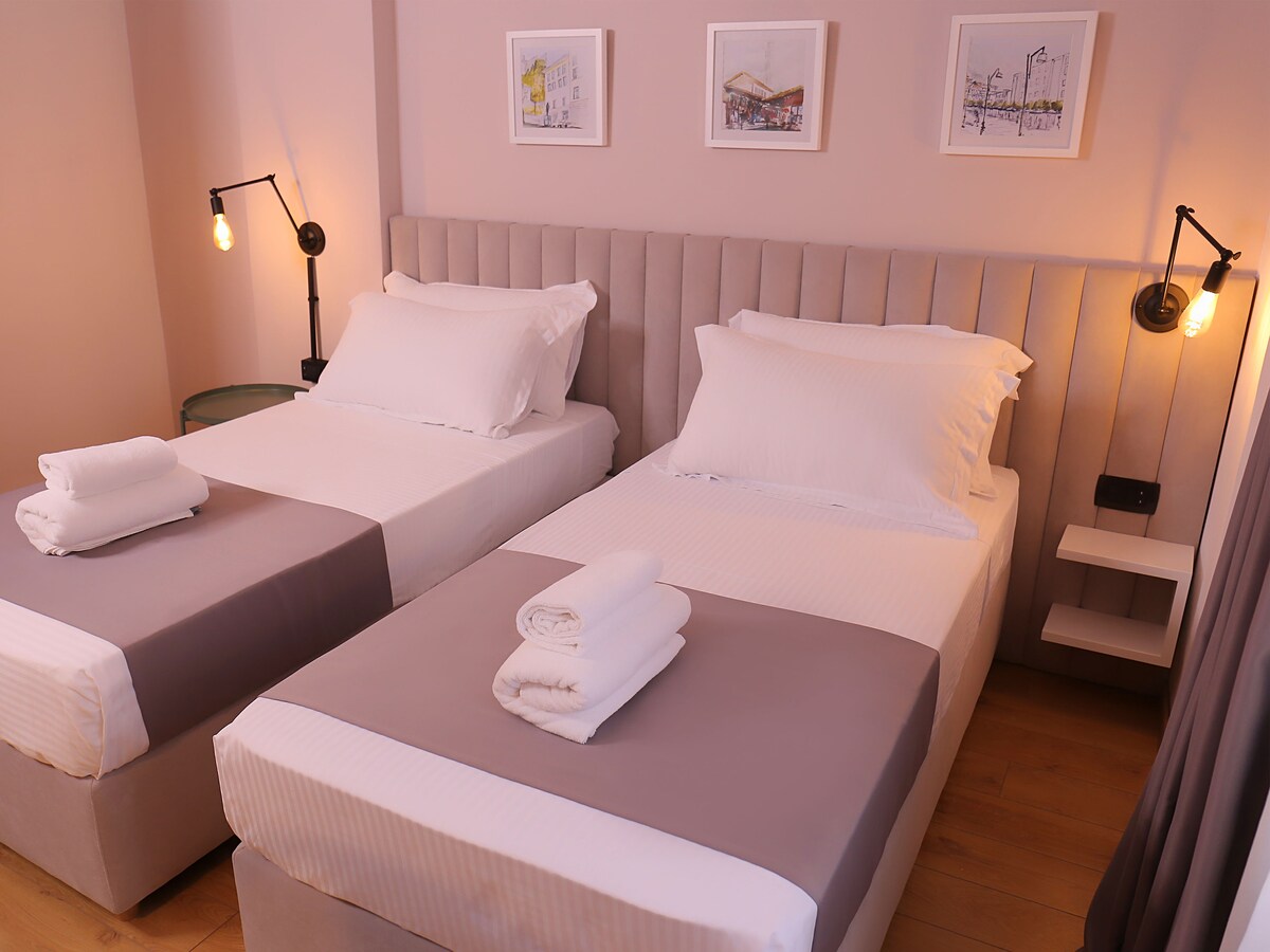 The sleeping area features two spacious beds with neutral-toned linens and neatly arranged white towels atop each. Minimalist bedside lighting enhances the soft atmosphere, while framed artwork is displayed above the headboard, adding a touch of character to the room.