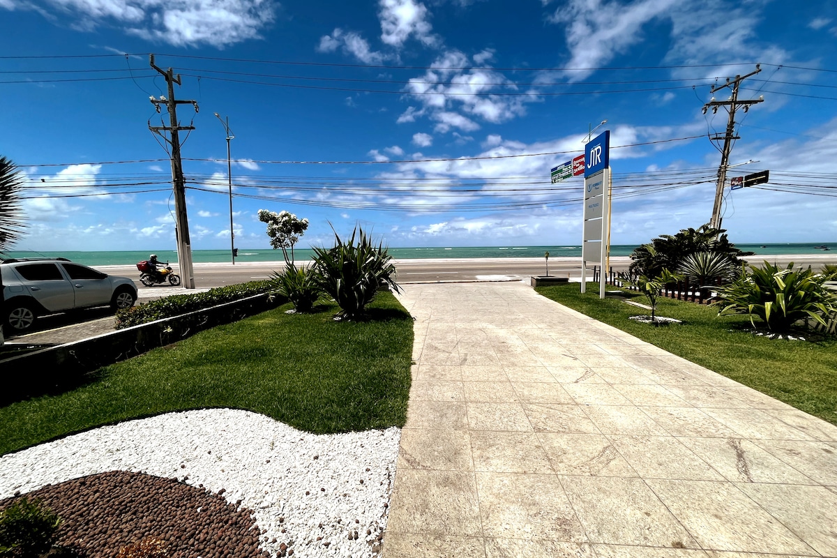 A scenic view from the property showcases well-maintained grass and decorative stones leading towards the road. The expanse of Jatiúca beach is visible beyond the street, with the ocean meeting the sky under a bright blue canopy dotted with clouds.