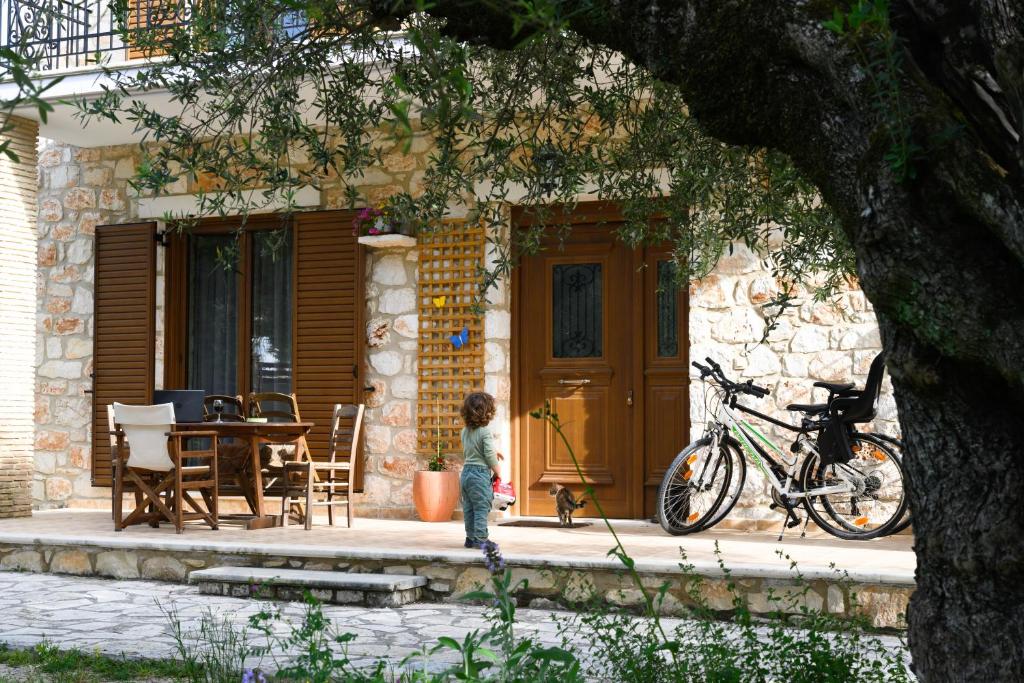 The entrance of the stone house is framed by olive trees, featuring a wooden door. A small patio area contains a wooden dining table with chairs, and two bicycles are parked nearby. Natural light highlights the stone facade, creating a serene outdoor setting.