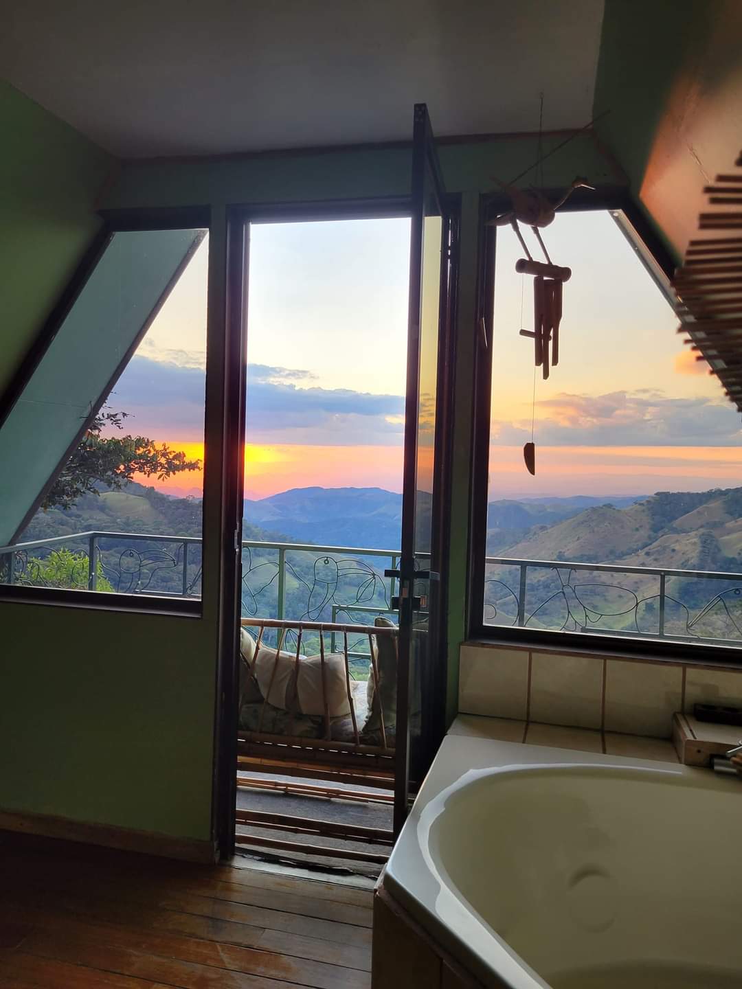 A doorway opens to a stunning sunset view over rolling mountains, framed by an A-frame structure. Light filters in, highlighting a whirlpool bathtub with a view in the foreground, while various decorative elements enhance the serene atmosphere.