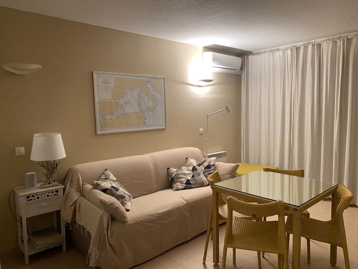 A light and airy studio space is presented with a comfortable sofa adorned with decorative pillows. A glass dining table is set with yellow chairs, while a wall-mounted map adds character. Natural light filters through sheer curtains, enhancing the room's openness.