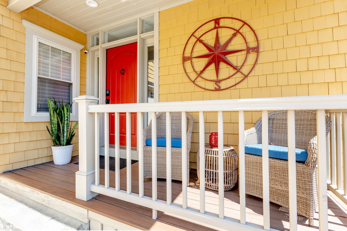 A welcoming porch is displayed, featuring two wicker chairs with blue cushions and a small round table. A vibrant red door serves as a focal point, while a decorative compass is mounted on the wall above. Lush greenery in a pot adds a touch of nature.