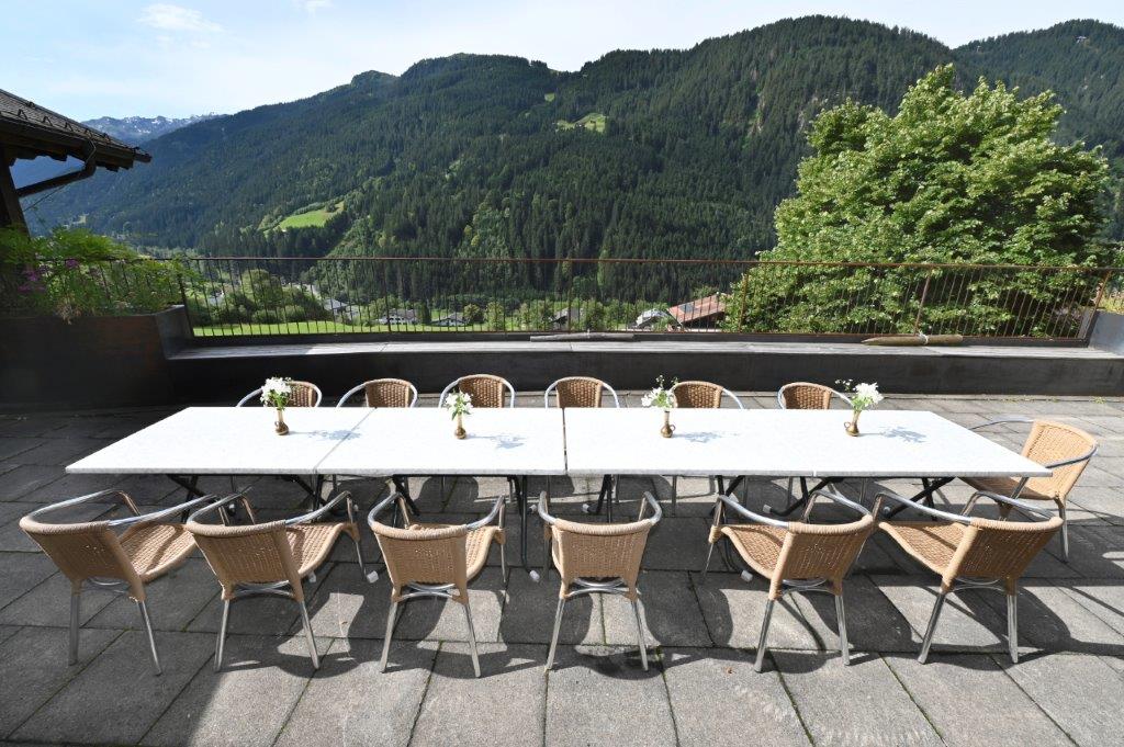 A large outdoor dining area features a long table surrounded by multiple wicker chairs. The backdrop showcases rolling green hills and mountains under a clear blue sky, creating a refreshing outdoor setting for gatherings.