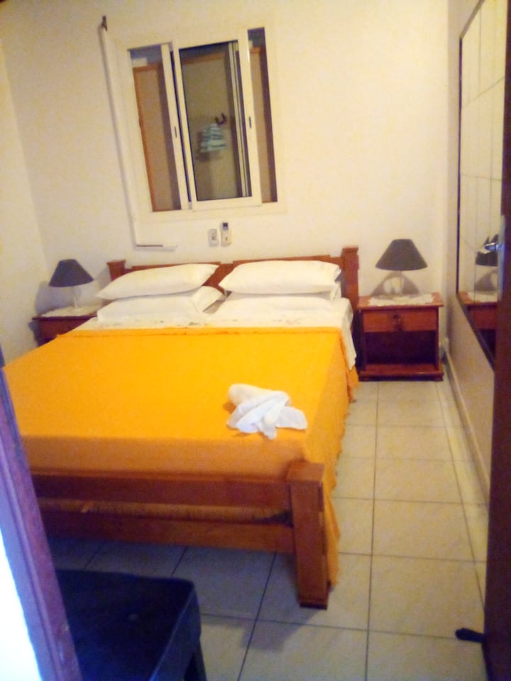 Тази стая е добре проветрена.  На лявата стена има прозорец като този, който виждате, гардероб и пейка зад леглото. 