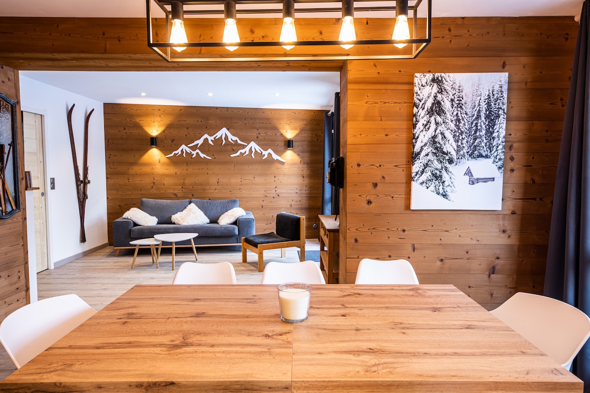 A spacious dining area features a wooden table surrounded by white chairs. In the background, a comfortable living space includes a gray sofa and coffee table, complemented by wooden wall accents and a mountain-themed artwork. Natural light brightens the cozy atmosphere.