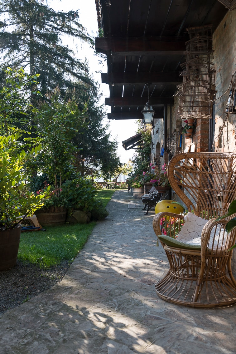 A stone pathway meanders through a lush garden, flanked by vibrant greenery and colorful flowers. Comfortable seating is provided with a rattan chair positioned nearby. The architectural features include wooden beams above, adding to the inviting ambiance of the outdoor space.