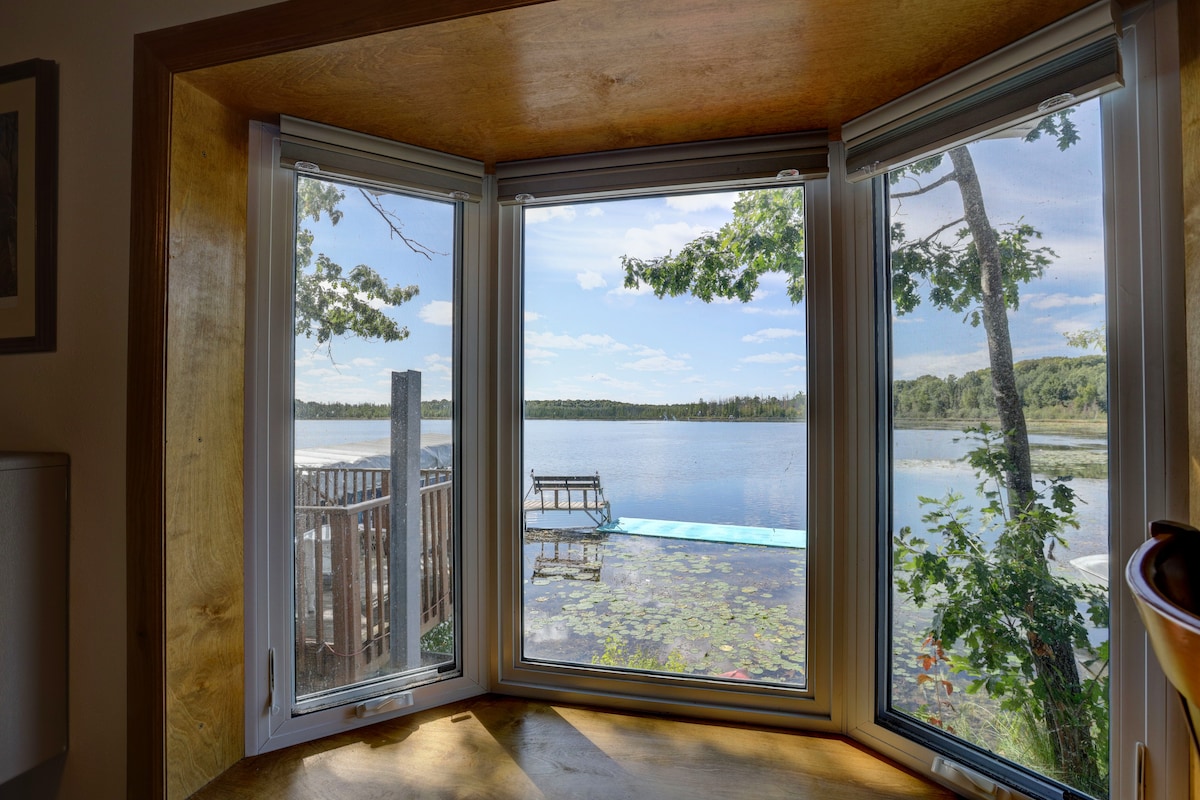 A multi-pane window offers a panoramic view of the serene Pickerel Lake, framed by lush greenery. The tranquil water reflects blue skies and gentle clouds, with a private dock visible in the foreground, surrounded by water lilies and natural vegetation.