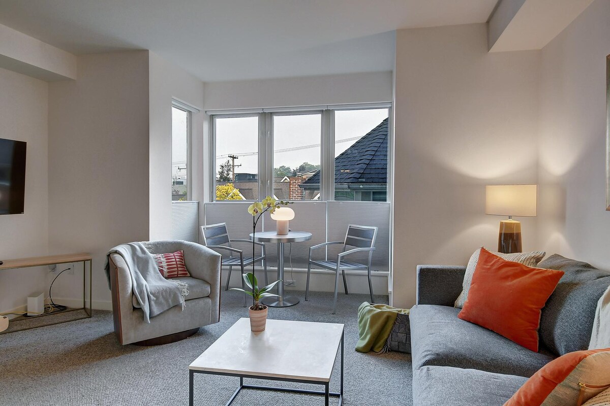 A bright living area features floor-to-ceiling windows, allowing natural light to fill the space. A comfortable sofa with orange and gray accents is complemented by a cozy armchair and a small dining table with two chairs. A potted plant adds a touch of greenery.