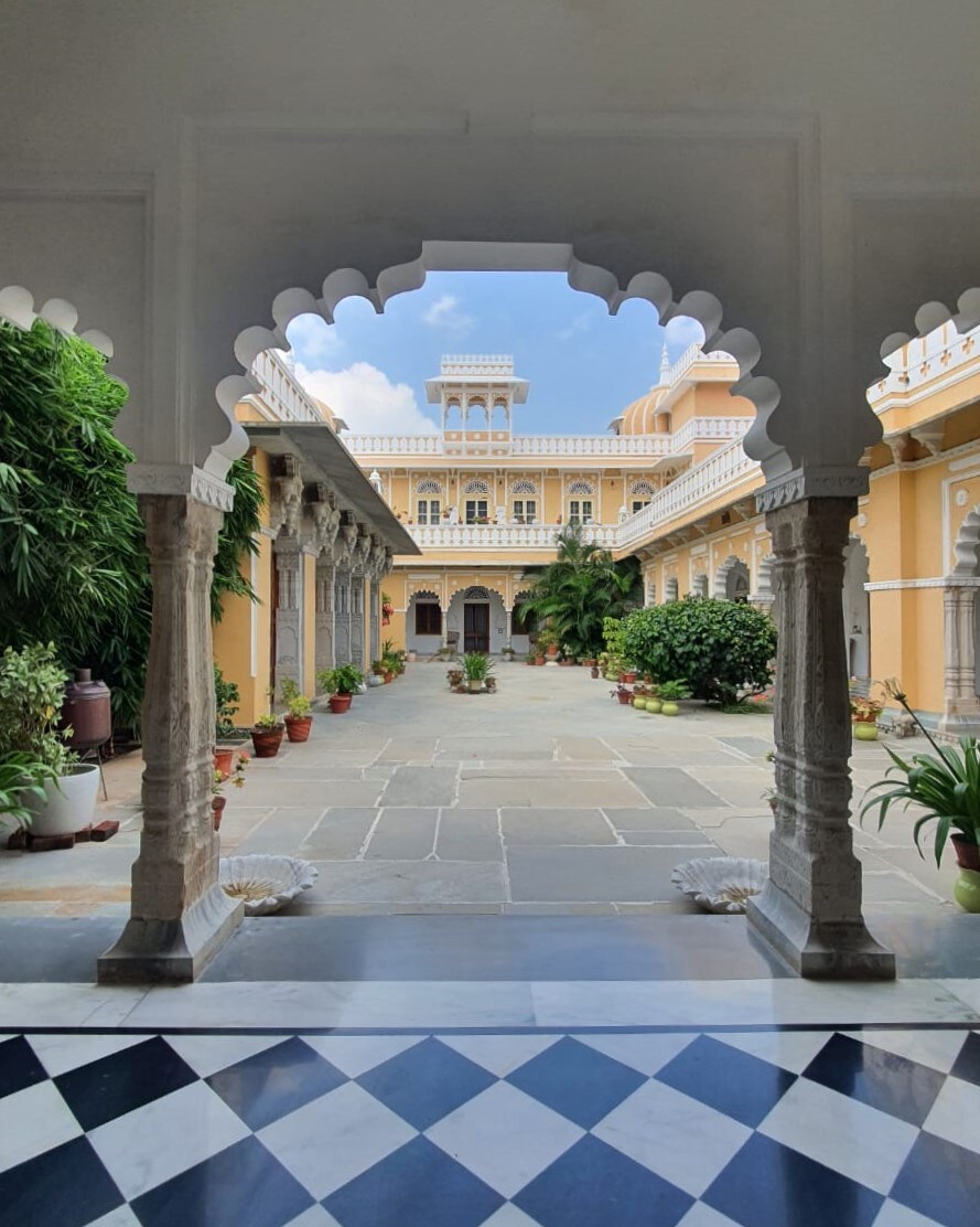 An archway with intricate carvings frames a serene courtyard filled with potted plants and lush greenery. The pale yellow walls of the surrounding structure rise above, showcasing ornate architectural details and an inviting atmosphere, bathed in natural light from the open sky.