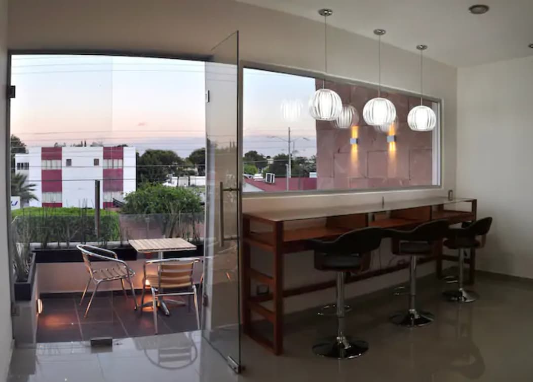 A modern workspace features a glass wall that provides a view of the outside area. Stylish pendant lights hang above a wooden counter with bar stools. A small balcony area holds a seating arrangement, and the space is illuminated by natural light.