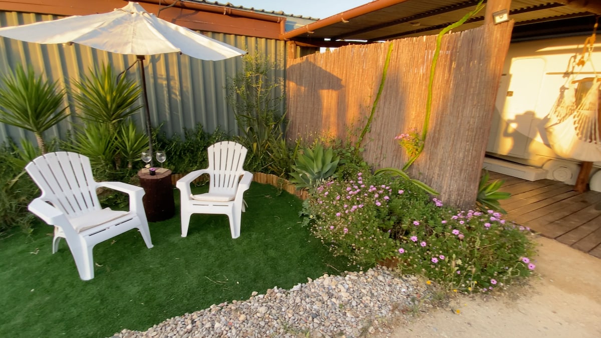 A private garden area is presented with two white lounge chairs positioned on artificial grass. A small table sits between the chairs, and a large umbrella provides shade. Lush greenery and vibrant flowers enhance the tranquil atmosphere, with a wooden structure visible in the background.