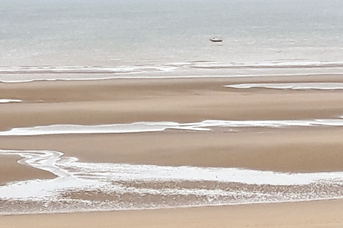 A serene view of the beach showcases soft, sandy shores meeting gentle waves. Patterns of water meander across the dry sand, with a small boat visible in the distance, highlighting the tranquil coastal landscape.