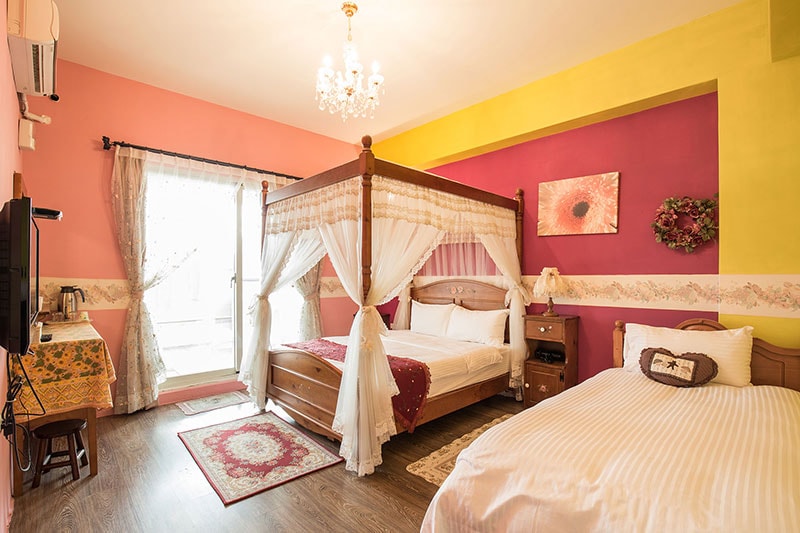 A bright bedroom features a four-poster bed draped with sheer fabric, complemented by a cozy twin bed nearby. Colorful walls in pink and yellow add vibrancy, while natural light enters through glass doors leading to an outdoor space.