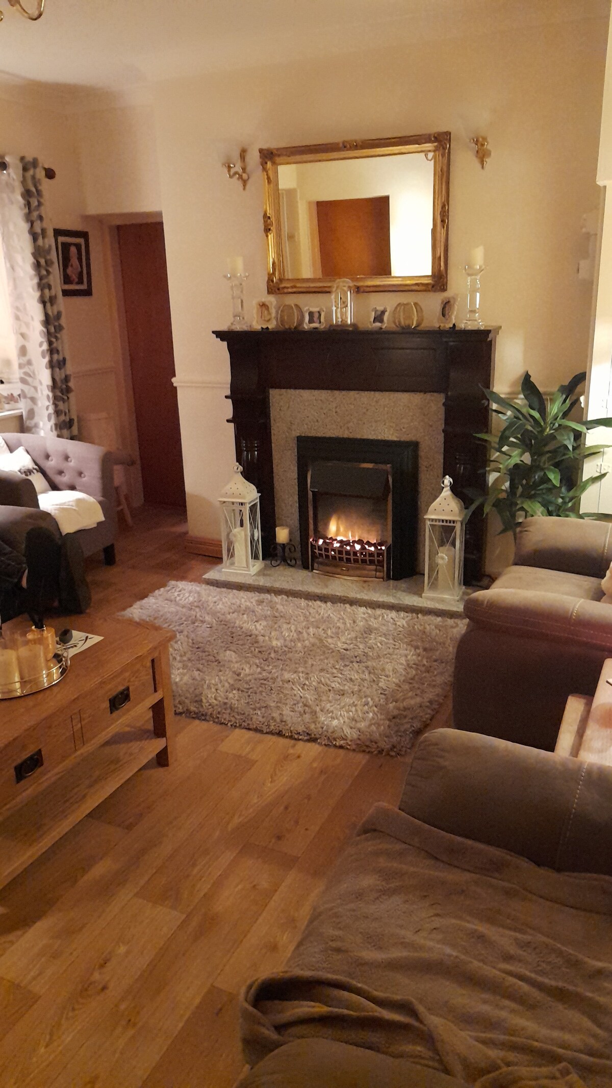 A warm living space features a decorative fireplace with a modern design, surrounded by two cozy seating areas. A rug lies on the wooden floor beneath a central coffee table, and a plant adds a touch of greenery. A mirror reflects soft lighting throughout the room.