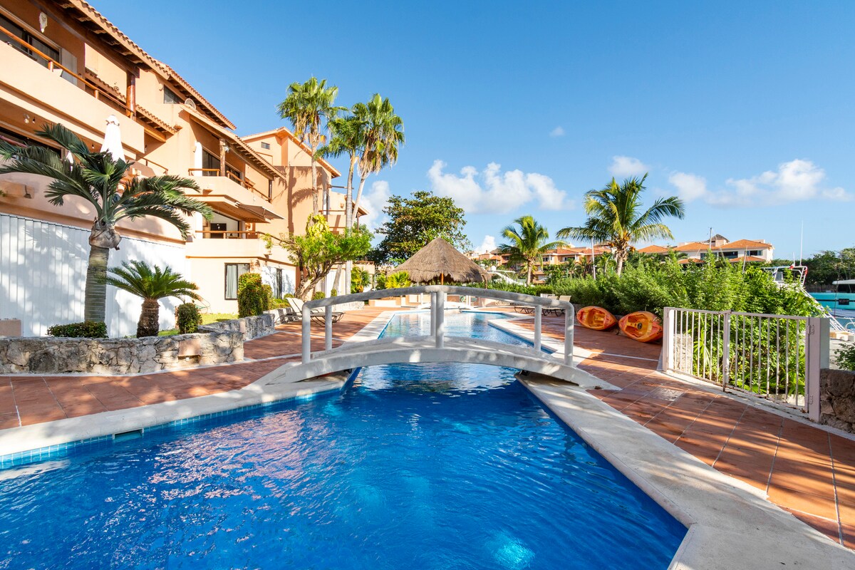 A serene swimming pool is framed by vibrant palm trees and lush greenery. A small bridge spans the pool, leading towards a thatched-roof structure. Sunlight reflects off the water, highlighting the inviting blue tones of the pool area.