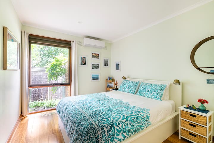 A cozy bedroom with a large window overlooking a garden. The bed features a turquoise and white patterned duvet and matching pillows. The room has wooden floors, a round mirror on the wall, and a small bedside table with drawers and a vase of flowers.