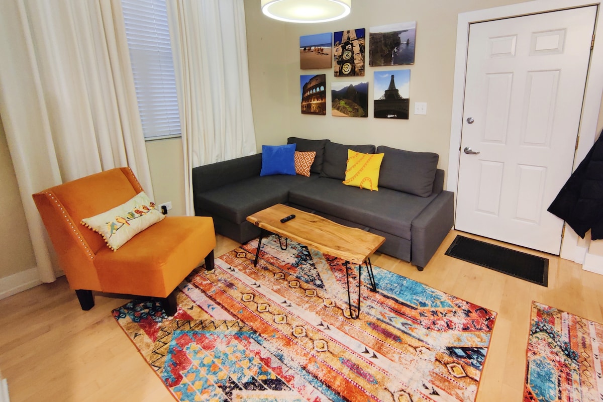 A cozy living area features a modern sectional sofa adorned with colorful cushions and a vibrant area rug. A wooden coffee table provides a natural accent, while bright artwork is displayed on the walls. Natural light filters through the window, and the main entrance is visible.