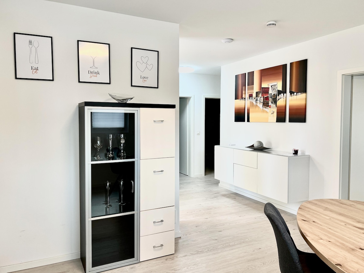 A modern living area features a stylish sideboard with glass doors showcasing elegant drinkware. Above, framed wall art displays themes of eating, drinking, and love. A sleek dining table is positioned nearby against a neutral-toned wall, contributing to the overall contemporary design.