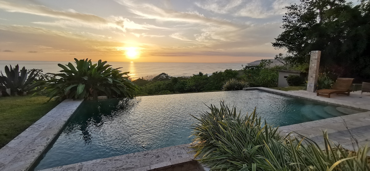 An infinity pool reflects the vibrant sunset over the ocean, surrounded by lush greenery. The scene features a serene landscape that transitions from water to grass, offering a peaceful retreat. A few lounge chairs are positioned nearby for relaxation.