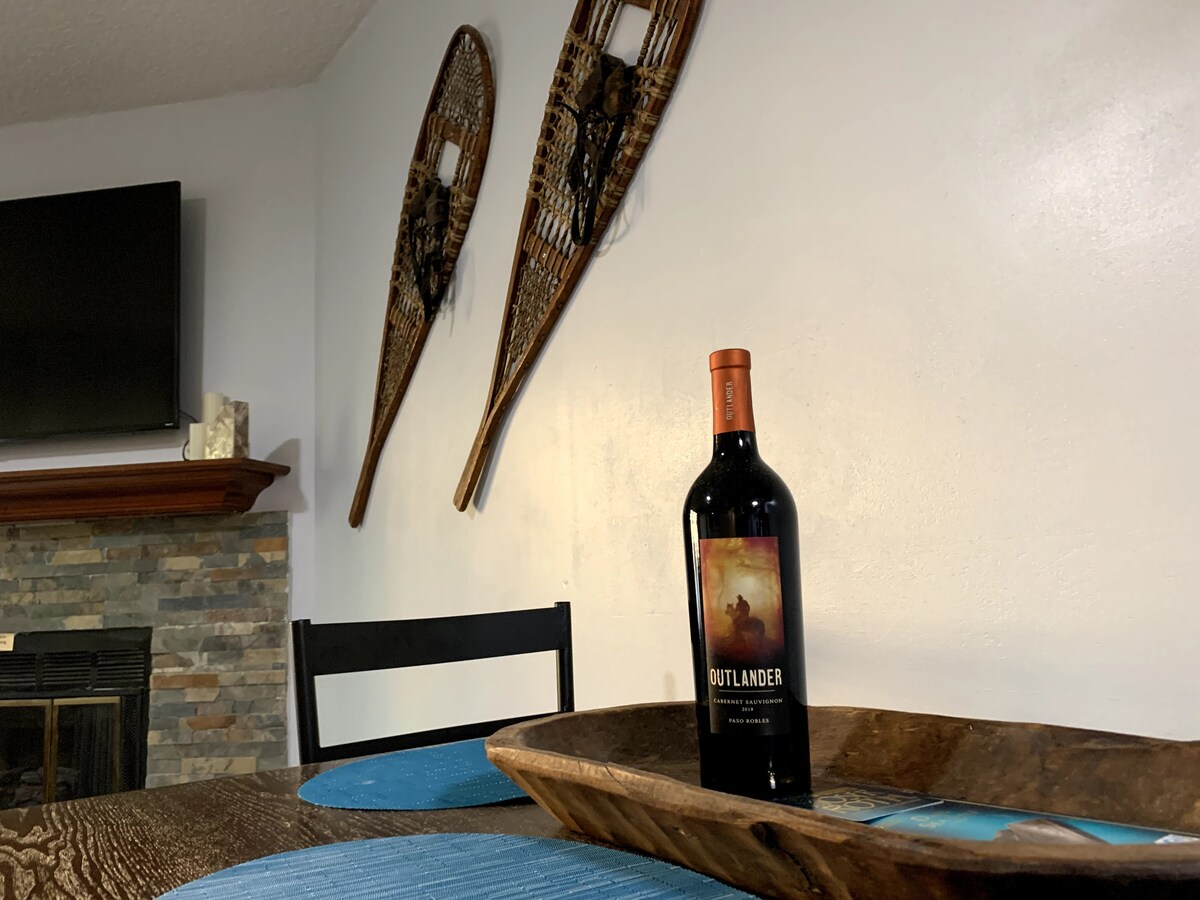 A wooden tray holds a bottle of wine, positioned on a dining table near a gas fireplace. Two decorative snowshoes are mounted on the wall, complemented by a television above the mantel. Blue placemats add a pop of color to the table setting.