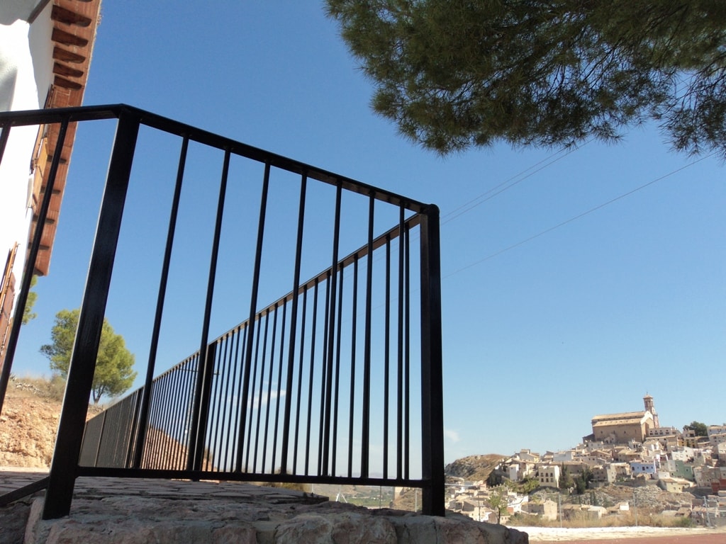 A black metal railing is visible in the foreground, overlooking a scenic view of the distant village set against a clear blue sky. The terrain is rocky with sparse vegetation, hinting at the natural surroundings.