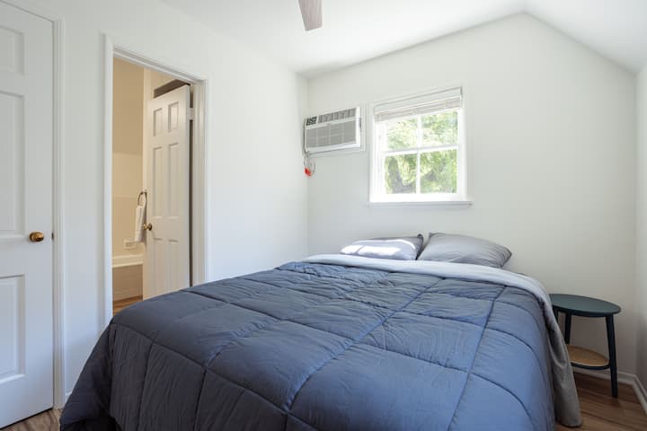 	- 	Dormitorio pequeño con ventana
	•	Cama tamaño queen con edredón
	•	Aire acondicionado sobre la ventana
	•	Pequeña mesita de noche redonda con la parte superior negra y patas de madera
	•	Abre la puerta que conduce a un baño.
	• 	Ambiente luminoso y acogedor
	•	Decoración minimalista