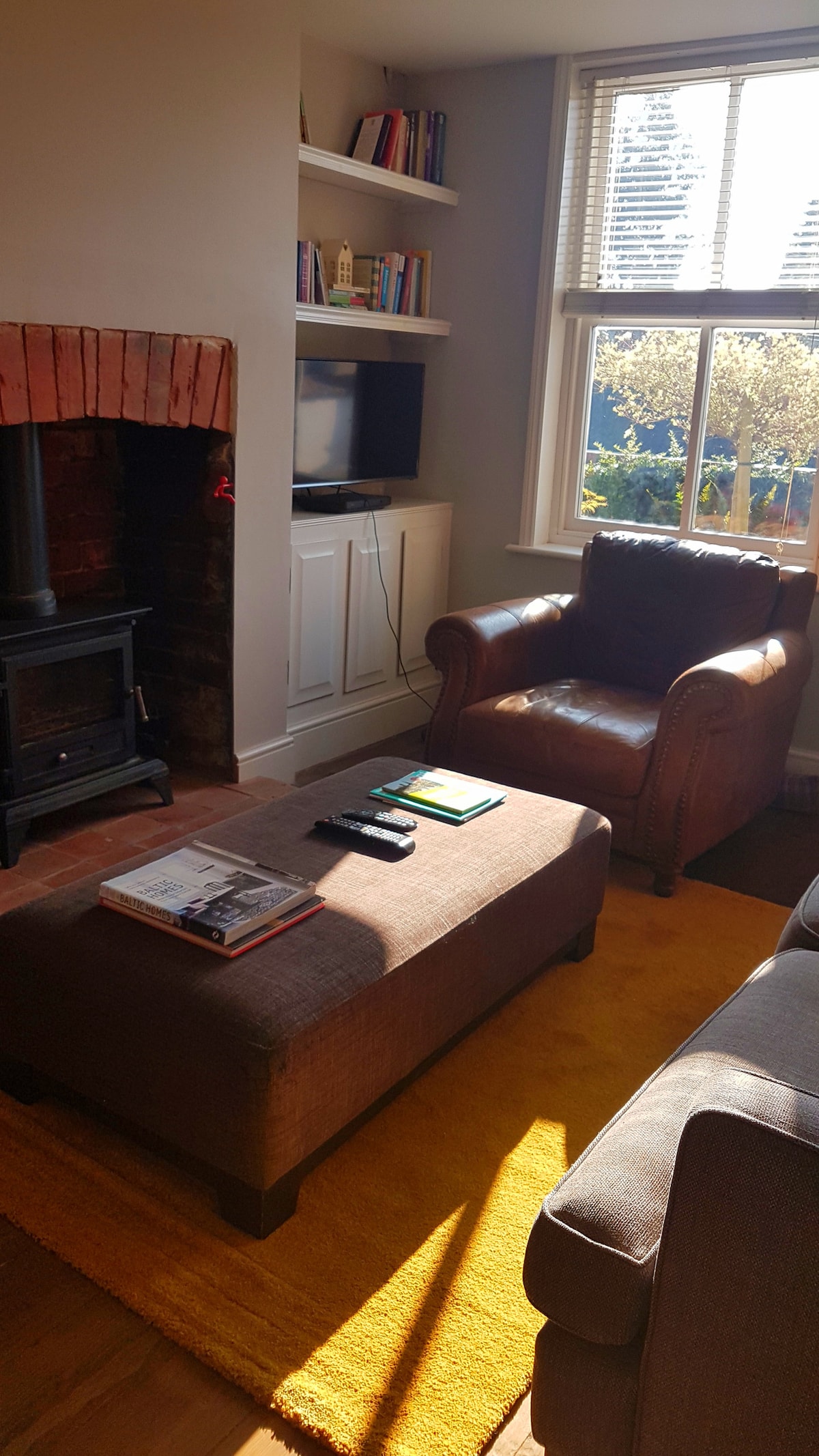 A warm living area features a rustic stone fireplace with a wood-burning stove. Comfortable leather armchairs and a plush ottoman are arranged on a cozy yellow rug. Sunlight filters through a window, illuminating bookshelves filled with books and adding a welcoming touch.