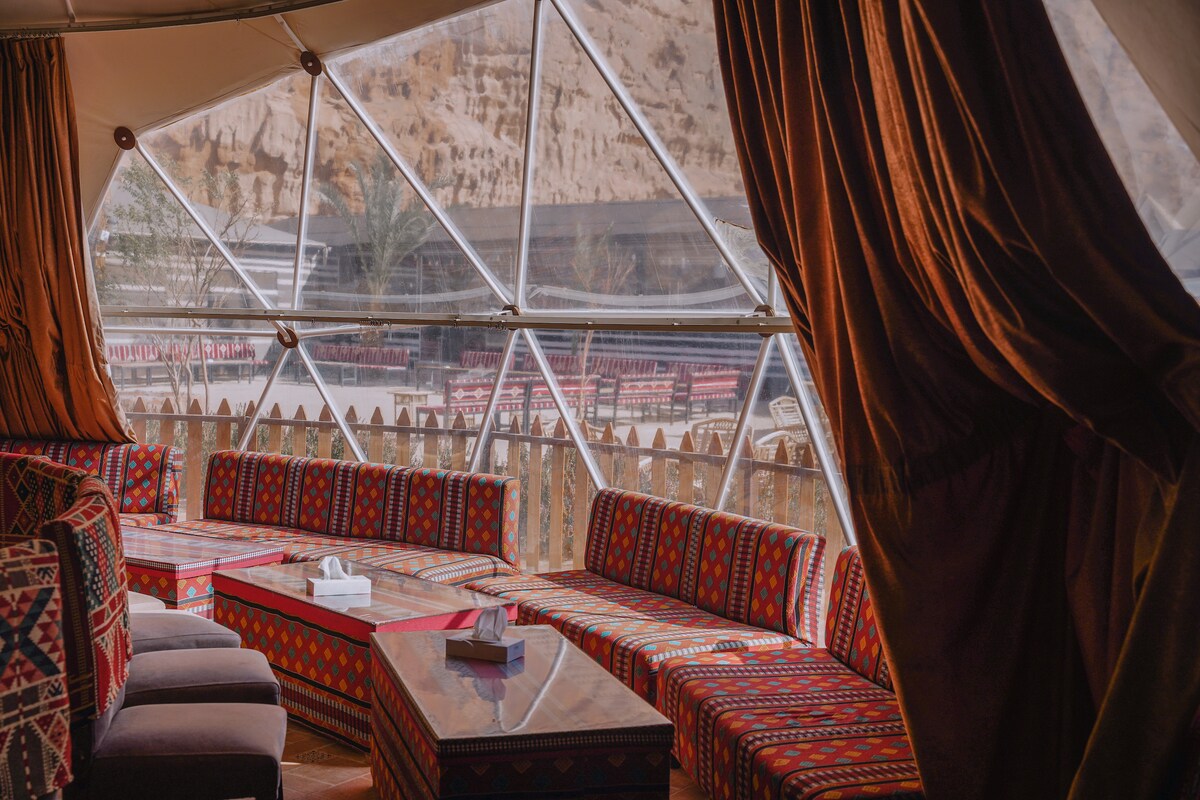 A spacious lounge area is framed by large windows, providing views of the desert landscape. Colorful fabric-covered seating lines the room, complemented by a low wooden table. Warm-toned curtains hang gracefully, enhancing the area's traditional ambiance.