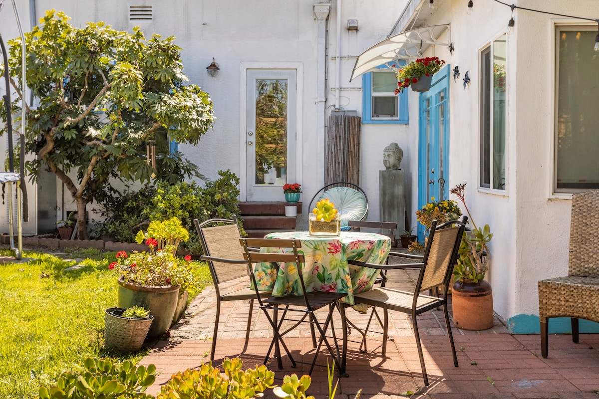 A garden patio is shown with a table surrounded by black chairs and adorned with a floral tablecloth. Various potted plants add greenery, while a wall with bright blue accents contrasts with the white exterior of the home. A doorway is visible, leading to the interior.