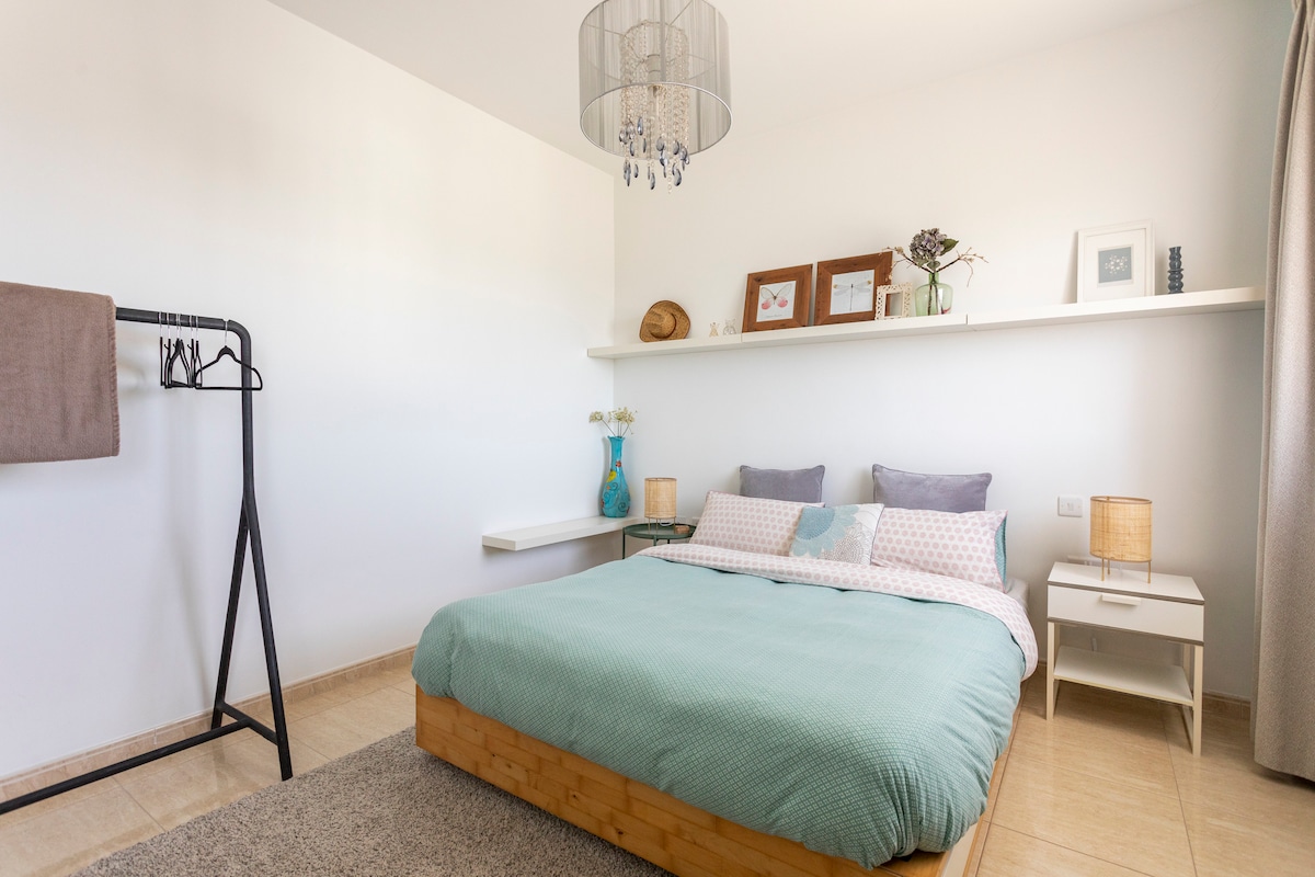 A spacious bedroom features a wooden bed with a soft, light blue coverlet. Framed artwork and a decorative shelf adorn the wall above the bed, complemented by a stylish chandelier. A modern clothing rack and bedside tables with small lamps complete the minimalist design.