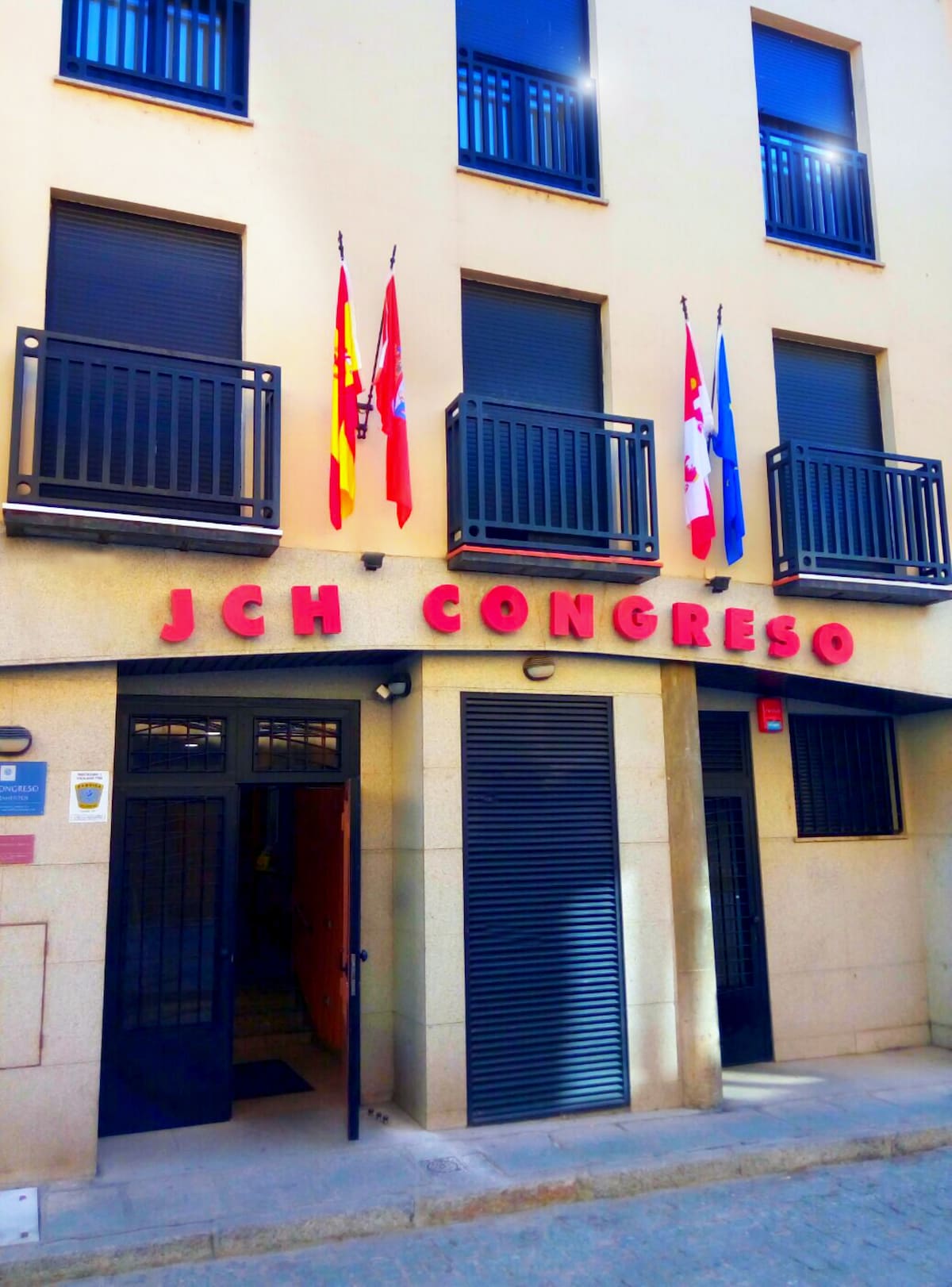 The entrance of the building is displayed, featuring the name 'JCH Congreso' prominently featured above the door. Several flags are neatly arranged above the entrance, and two balconies are visible on the upper floors, showcasing a modern facade.