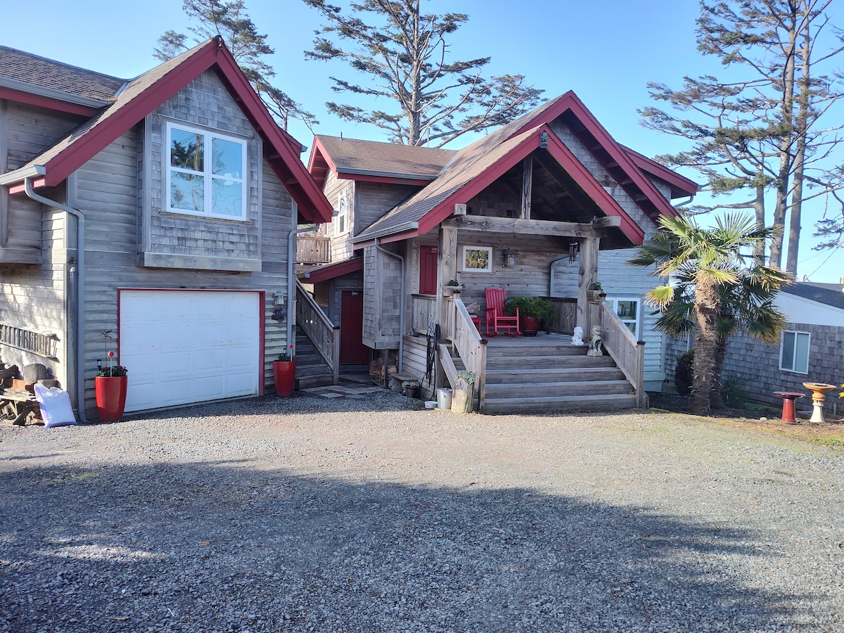The exterior of a two-story wooden building features a welcoming porch with red chairs. A gravel driveway leads to the entrance, with surrounding greenery and palm trees enhancing the natural setting. The architecture combines rustic and modern elements, reinforced by large windows.