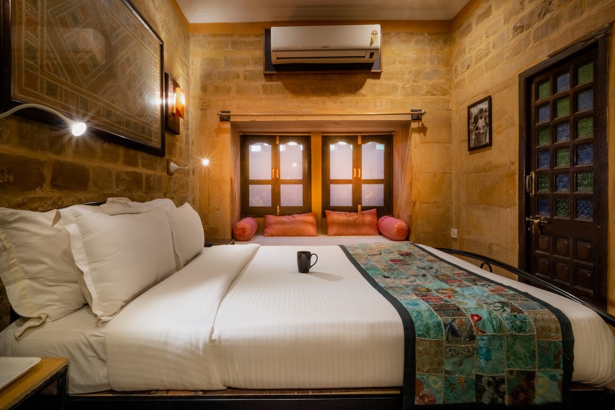 The Sandstone Room features a spacious bed adorned with a vibrant quilt and a decorative pillow. Soft lighting from bedside lamps enhances the rustic charm of the stone walls. A cozy seating area with plush cushions is visible through traditional wooden windows, offering a glimpse of relaxing décor.
