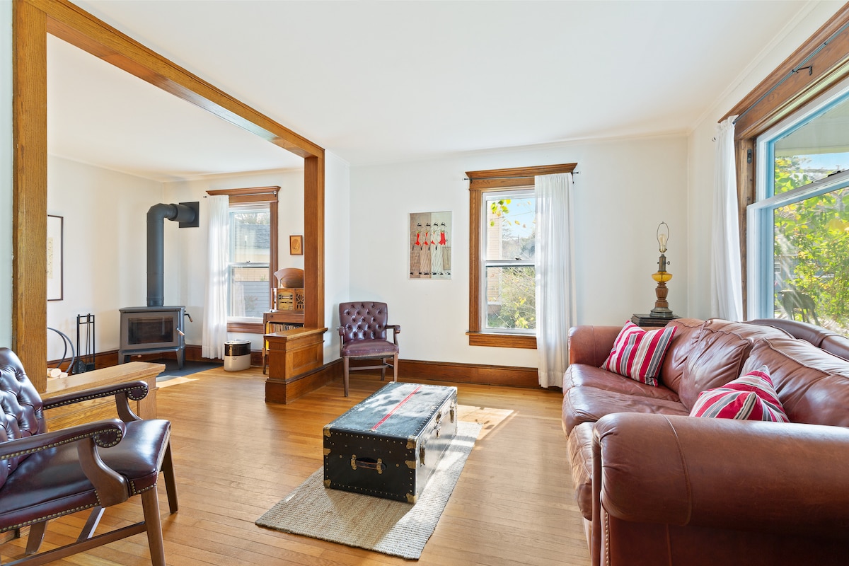 A spacious living room is highlighted by natural light filtering through large windows. A cozy leather couch and two armchairs provide seating, while a rustic wooden coffee table sits center stage. A wood-burning stove adds warmth, complemented by hardwood flooring and minimal decor.