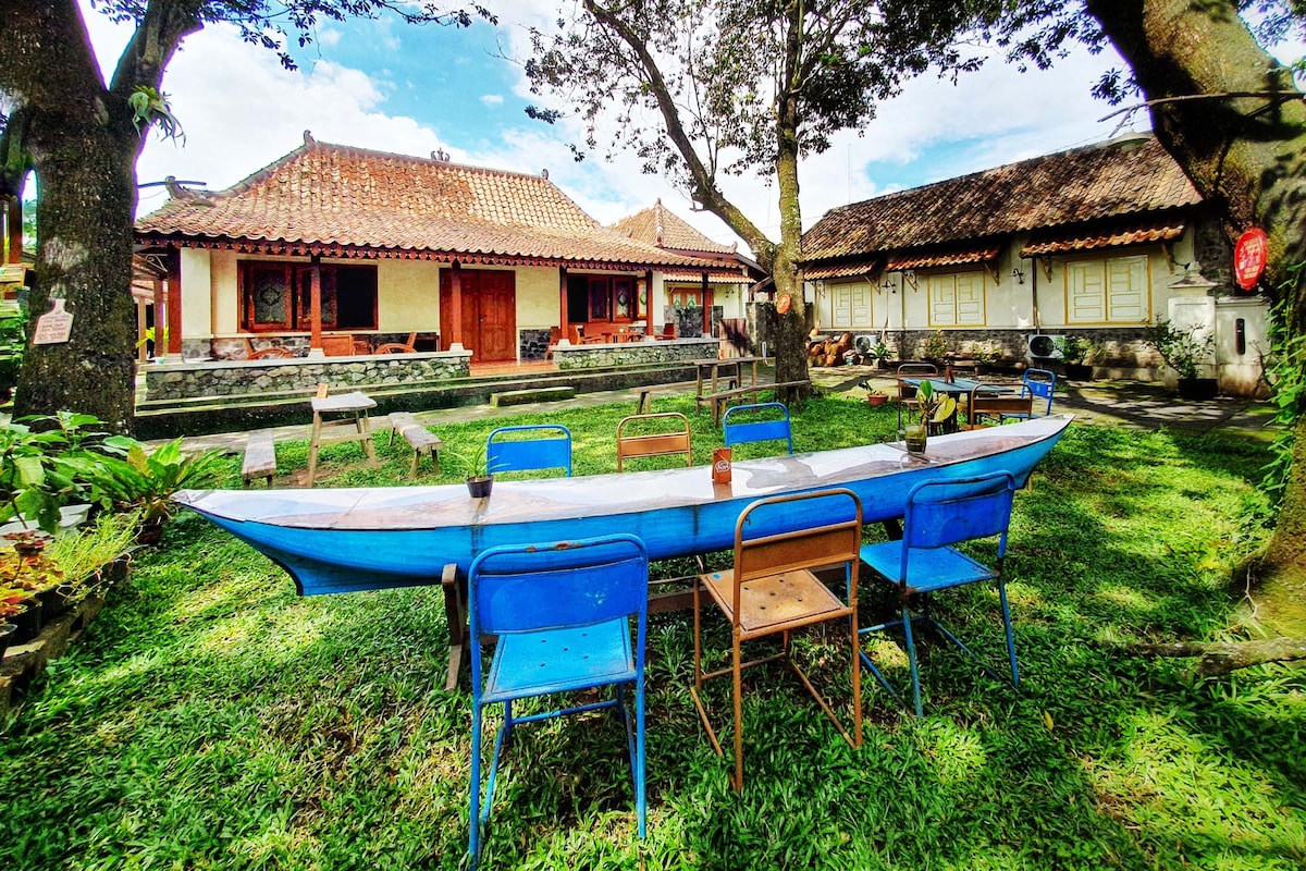 A spacious outdoor area is surrounded by trees, featuring a long blue table with several chairs placed around it. Traditional Javanese houses are visible in the background, showcasing wooden architecture and tiled roofs. Lush greenery creates a peaceful and inviting setting.