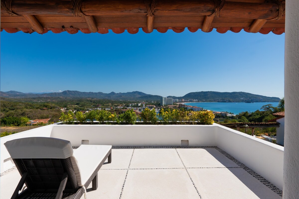 A spacious terrace features a sun lounger, offering panoramic views of the surrounding landscape, including lush greenery and distant mountains. The clear blue sky complements the tranquil atmosphere, while the horizon showcases a glimpse of the ocean.
