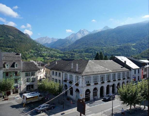 A panoramic view shows the charming village nestled in a valley, with majestic mountains in the background. The scenery includes trees lining the streets, and buildings typical of the region. Clear blue skies and soft clouds enhance the tranquil atmosphere.