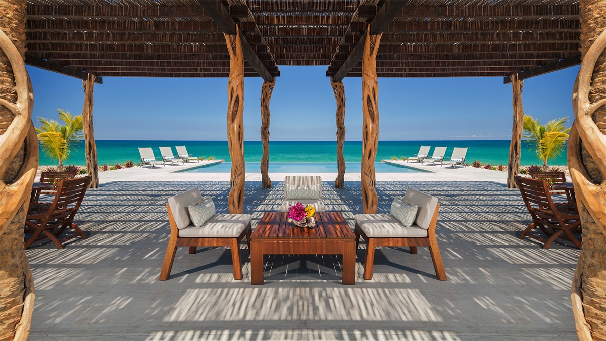 An outdoor seating area features wooden furniture with plush cushions, arranged around a low table adorned with a floral centerpiece. In the background, sun loungers are positioned by the turquoise sea, framed by palms and a clear blue sky.