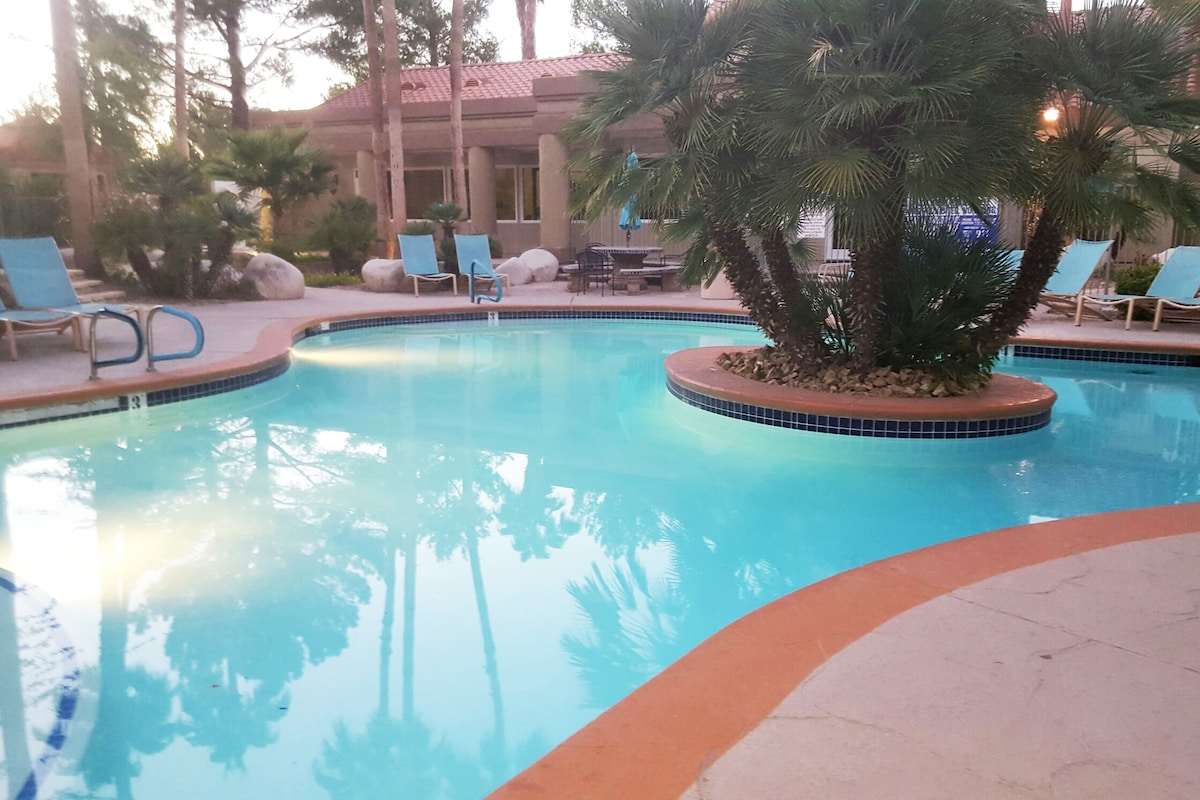 The community pool is surrounded by palm trees and comfortable lounge chairs. Clear water reflects the surrounding greenery, creating a relaxing environment. The area features a gentle slope from the poolside, inviting guests to enjoy the natural setting within the condominium complex.