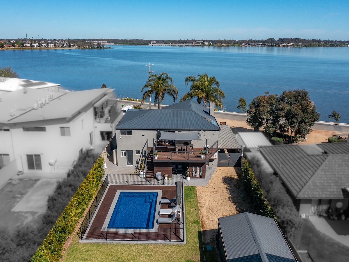 An aerial view captures a modern property with a spacious outdoor area featuring a swimming pool. Surrounding the pool are sun loungers and a lawn. The property overlooks Lake Mulwala, providing a scenic backdrop of water and trees.