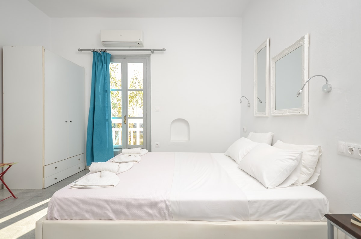 A bright and airy bedroom features a spacious bed with crisp white linens. Light pours in through a window adorned with light blue curtains. Two mirrors are mounted above bedside fixtures, and a wardrobe is visible against the wall, contributing to the room's minimalist decoration.
