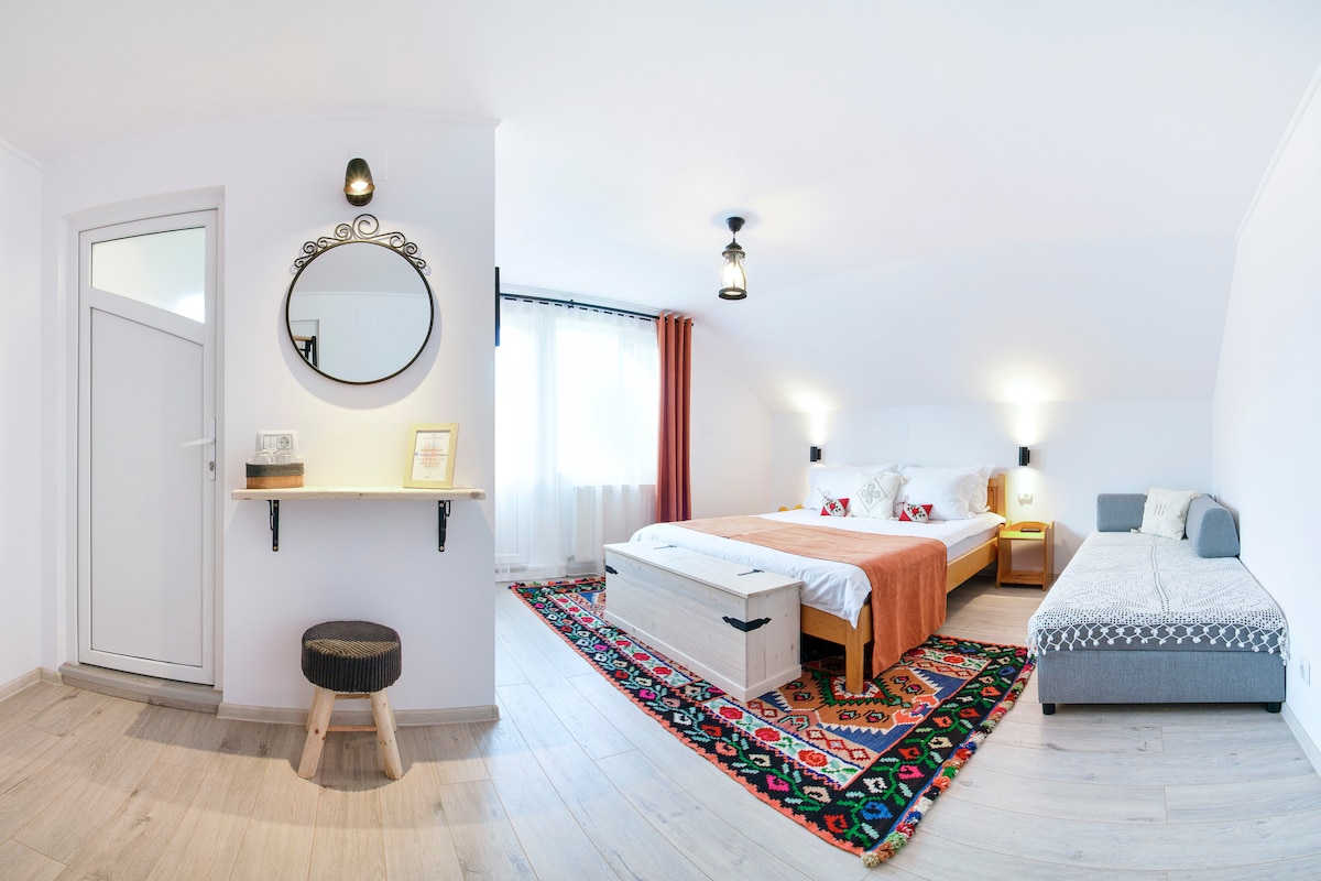 A spacious bedroom features a light color palette, with a large bed complemented by a cozy throw. A seating area with a gray upholstered chair is present. Natural light floods in through the window, enhancing the room's inviting atmosphere. A round mirror and a small shelf add functional decor.
